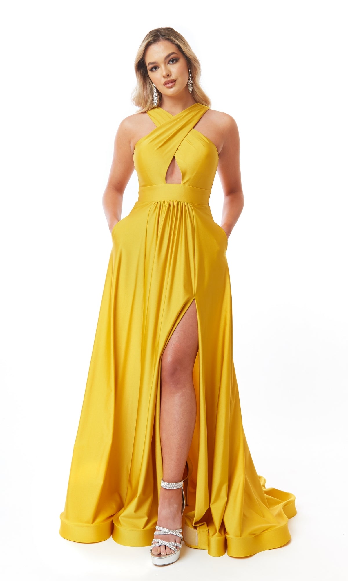 Woman in the Atria 6706H Cut-Out Long A-Line Prom Dress, featuring a yellow floor-length design with a front slit and crisscross halter neckline, posing against a white background.